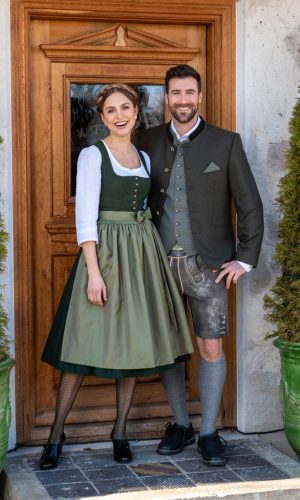 Trachtenpaar - Paarbild Frau mit grünem Dirndl, weißer langarm Bluse und Herr mit grünem Gilet weißes Hemd, bräunliches Sakko und grauer Lederhose (kurz) vor Eingangstür