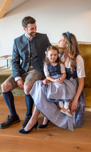 Familienbild auf Couch - Herr mit Sakko und Gilet, grauer Lederhose und blauen Stutzen, Dame mit blauem Dirndlkleid gemustert und hellblauer Schürze und Kind im Dirndl blau gemustert und hellblauer Schürze