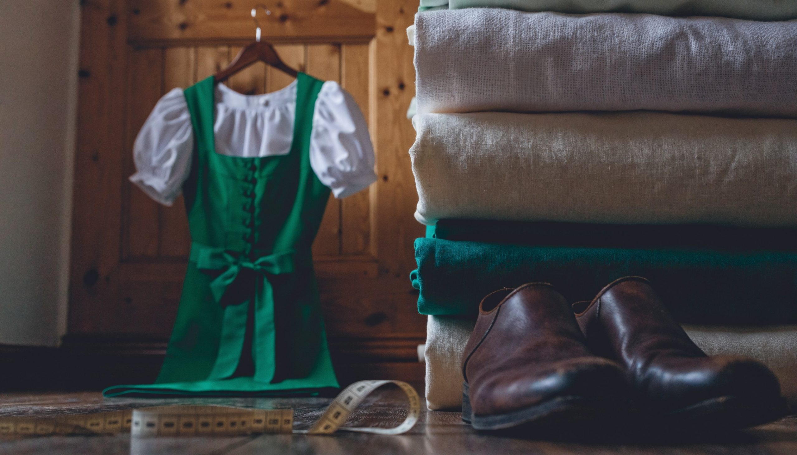 Dirndl kaufen: Dirndl mit Schürze und weißer Bluse auf einem Kleiderbügel, daneben Stoffe und Trachtenschuhe