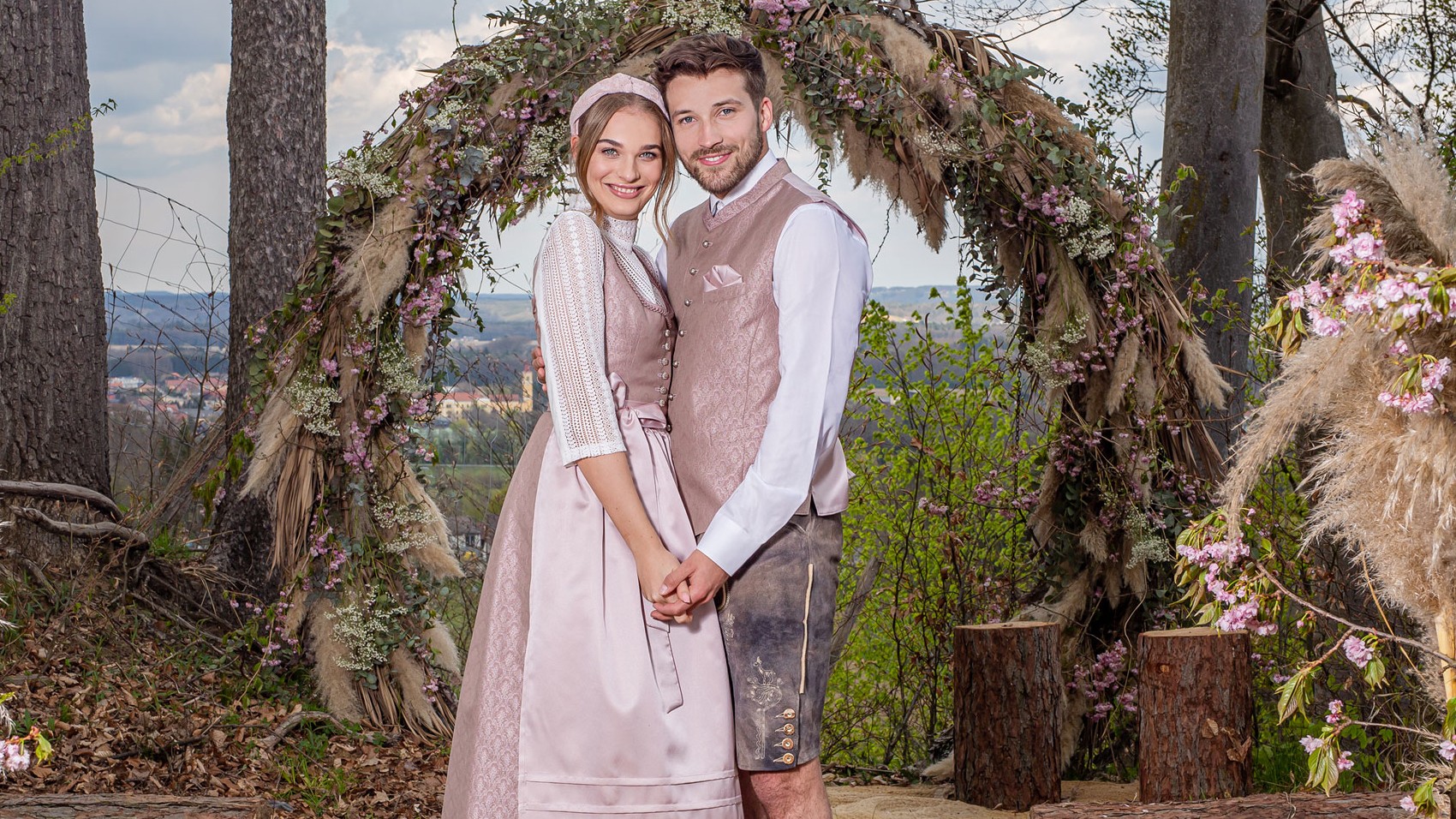 Hochzeit in Tracht: Brautpaar in rosé Dirndl und Lederhose mit Gilet unter einem Blumenbogen