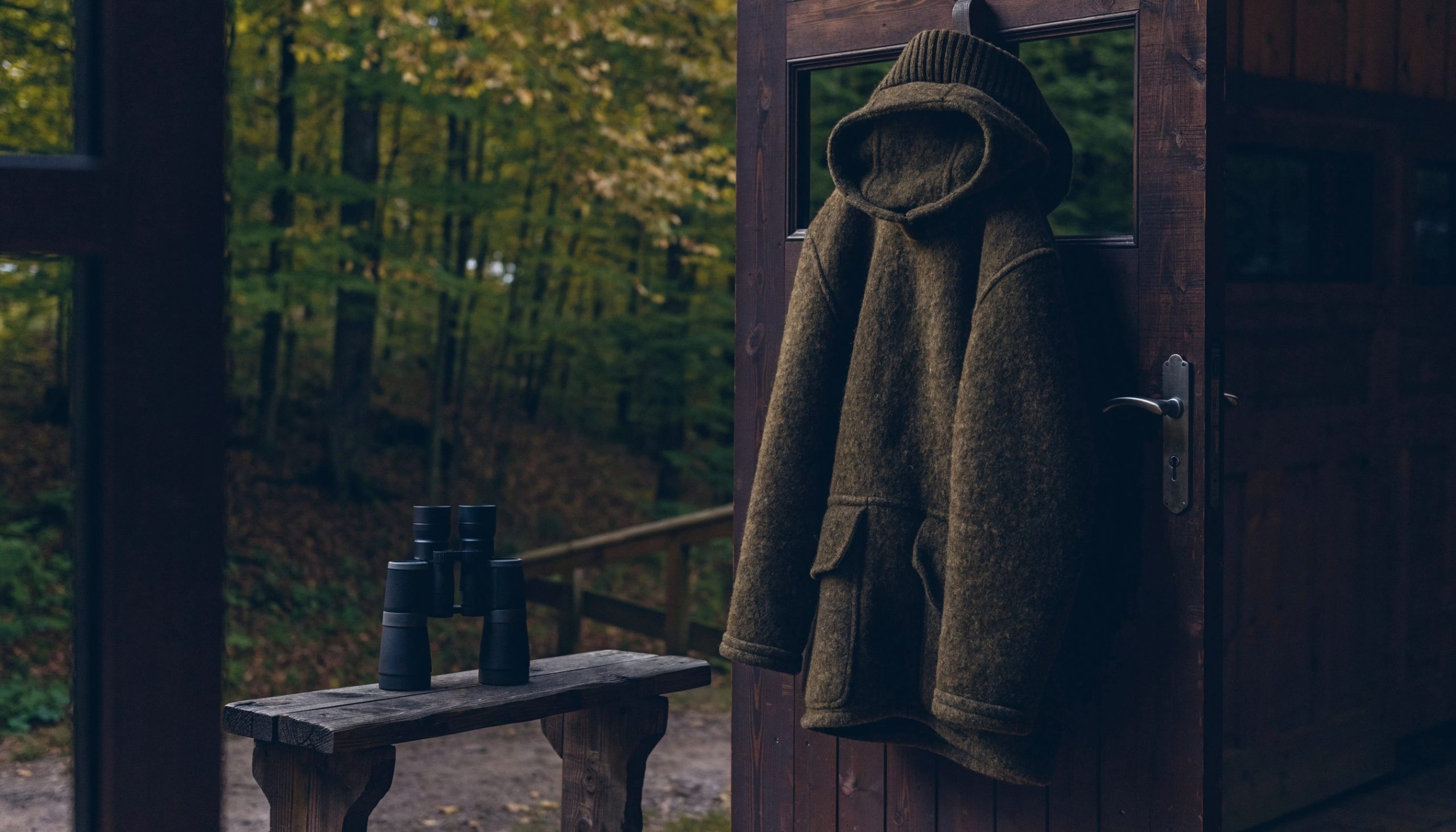 Lodenjacke für Herren mit Kapuze hängt an der Tür einer Jagdhütte, daneben ein Fernglas auf einer Holzbank