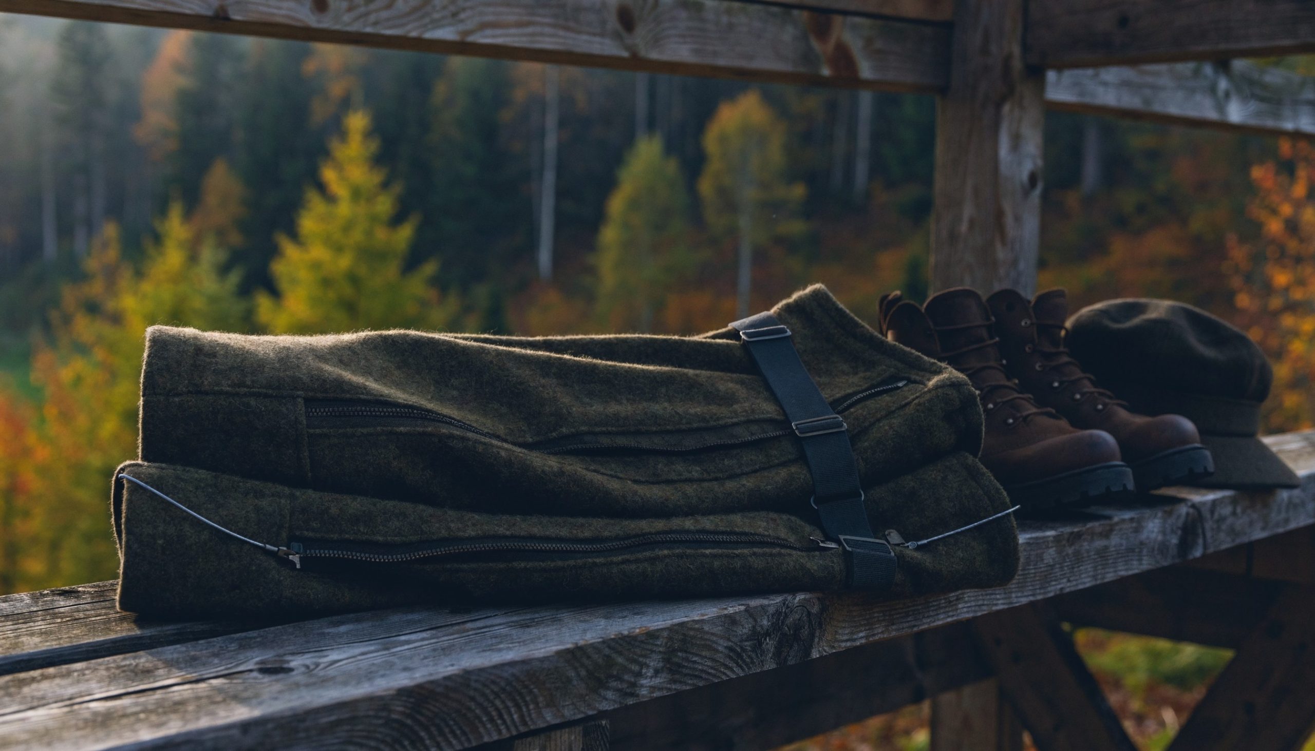 Multifunktionshose für Herren aus Loden mit Hosenträgern und Gamasche auf der Bank eines Hochstands, daneben Bergschuhe